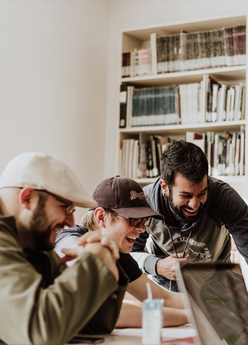 Trois jeunes hommes travaillant ensemble à une table, concentrés sur un ordinateur portable, dans une ambiance conviviale et studieuse avec des étagères de livres en arrière-plan.
