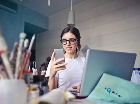 Jeune femme à lunettes travaillant sur ordinateur portable et consultant son téléphone, dans un bureau lumineux, illustrant l’agilité et la connexion des équipes Calidius.