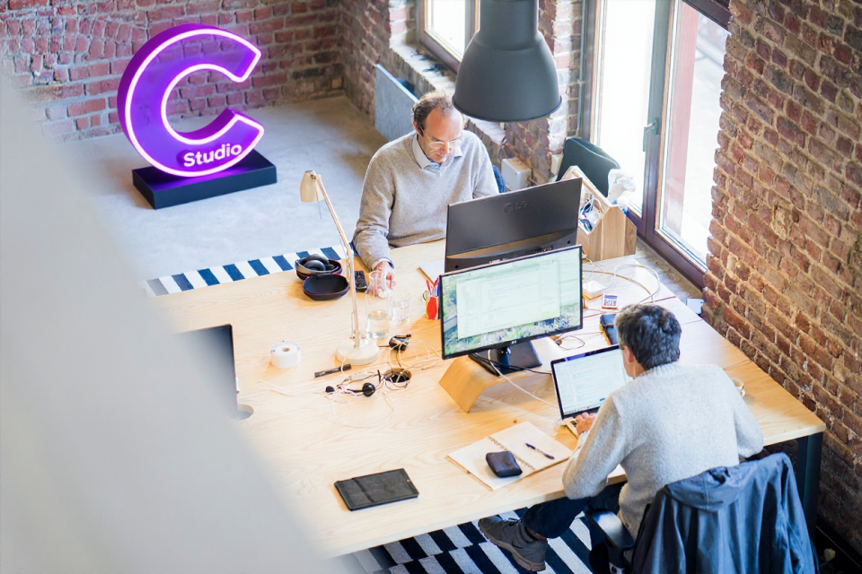 Deux personnes travaillant sur des ordinateurs dans un studio lumineux à murs de briques, avec un néon violet “C Studio”, illustrant l’ambiance collaborative et créative de Calidius Studio.