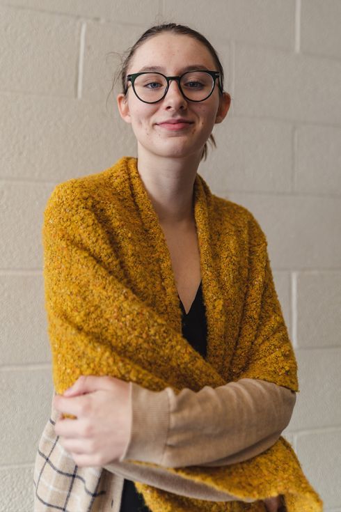 Jeune femme à lunettes, debout, souriante et enveloppée dans un large châle jaune moutarde, illustrant la bienveillance et l’authenticité au sein de la communauté Calidius.