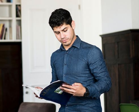 Jeune homme concentré lisant un carnet, debout dans un espace de travail moderne, illustrant la réflexion et la préparation au sein d’une équipe.
