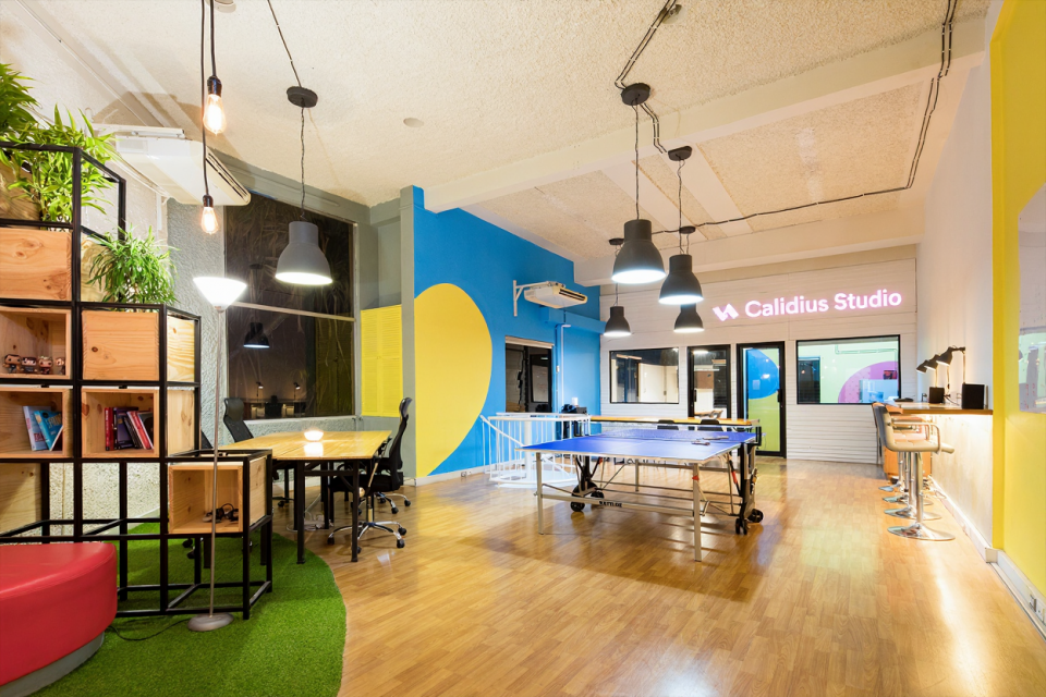 Vue d’ensemble d’un espace de coworking coloré avec tables de travail, plantes vertes, terrain de ping-pong et enseigne lumineuse “Calidius Studio”, illustrant la créativité et la convivialité du studio.