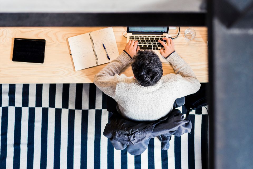 Vue plongeante d’une personne travaillant sur un ordinateur portable, avec carnet et tablette sur un bureau en bois clair, dans un espace de coworking lumineux.